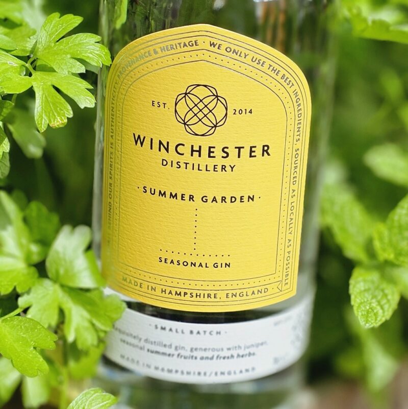 Close-up of a yellow-labeled Winchester Distillery Summer Garden Seasonal Gin bottle, surrounded by green herbs.