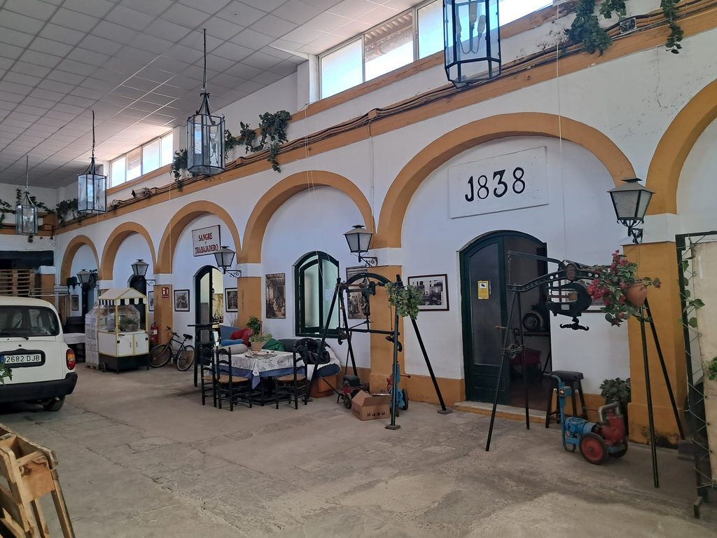 Wide interior courtyard shot showing arched arcade with "1838" on the wall, vintage winery equipment, iron lanterns and vine garlands