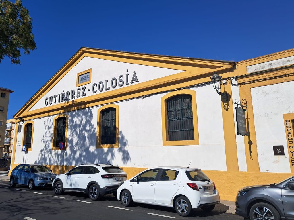 Exterior building shot — white and gold facade with "Gutiérrez-Colosía 1838" on the pediment, blue sky